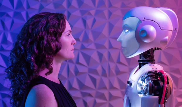 A woman and a humanoid robot face each other, illuminated by purple and blue light