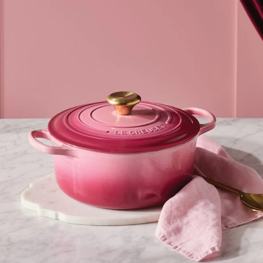 A Le Creuset Signature Round Dutch Oven in Berry color, with a gold knob, placed on a white marble trivet.